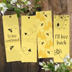 three tags with bees on them hanging from a wooden table next to white flowers and greenery