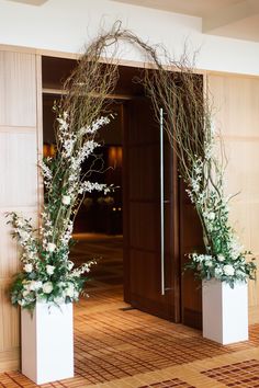 two tall white vases with flowers and branches on the floor in front of an open door