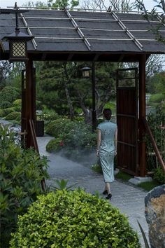 a woman walking down a path under a gazebo next to bushes and trees in the background