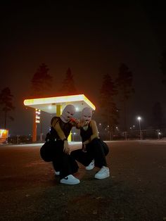 two people with masks on their faces sitting in front of a gas station at night