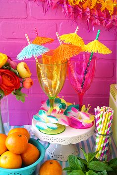 a table topped with fruit and drinks on top of a white cake stand next to a pink brick wall