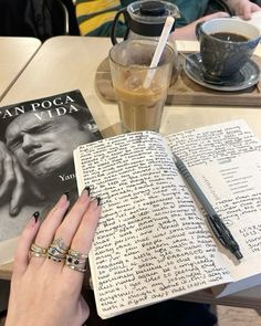 a woman's hand is holding an open book with writing on it and a cup of coffee in the background