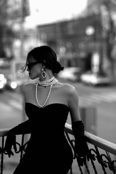 a woman in a black dress and pearls is leaning on a railing looking at the street