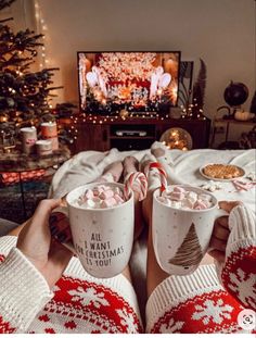 two people holding mugs with hot chocolate and marshmallows in front of a christmas tree