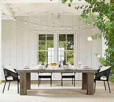 an outdoor dining table with lights strung from the ceiling and chairs around it in front of a white house