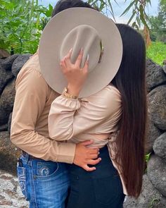 two people are hugging each other in front of some rocks and trees, one is wearing a cowboy hat