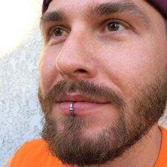 a close up of a man with a beard and piercing on his nose wearing an orange shirt