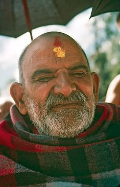 an old man with a yellow flower on his forehead is looking at the camera while standing under an umbrella