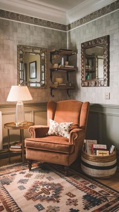 A cozy reading nook. A burnt-orange velvet armchair with a white floral cushion sits centrally, accompanied by a round wooden side table holding a table lamp with a pleated shade. Above the chair, there's a decorative mirror framed with intricate metalwork. Adjacent to the chair, on the wall, is a shelving unit displaying various decorative items. The floor is adorned with a multicolored rug, and a basket filled with books sits beside the chair. The room has a muted color palette with grayish wallpaper and a greenish wainscoting. Vintage Reading Chair, Vintage Reading Corner, Grayish Wallpaper, Reading Corner Home, Corner Home Decor, Round Wooden Side Table, Multicolored Rug, Vintage Armchair, Mirror Framed