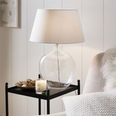 a glass table with a candle on it next to a white chair and a lamp