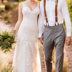 a bride and groom holding hands walking through the woods in their wedding attire with suspenders