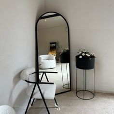 a mirror sitting on the wall next to a white chair and table with flowers in it