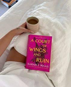 a woman laying in bed with a cup of coffee and book on her lap, reading a court of wings and ruin by sarah maas