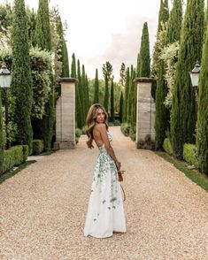 a woman in a white and green dress standing on a gravel road with trees behind her