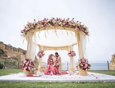 two people sitting on a white cloth covered stage with flowers and greenery around it