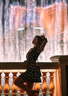 a woman in a black dress standing on a balcony next to a fountain at night