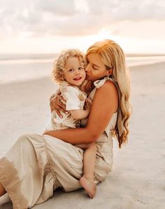 a woman holding a baby on the beach at sunset with clouds in the sky behind her