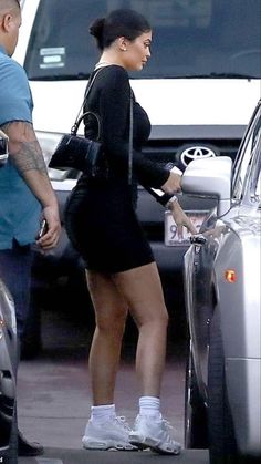 a woman in black dress standing next to a car with her hand on the door handle