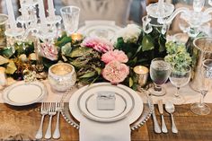 the table is set with white plates and silverware, pink flowers and greenery