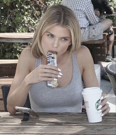 a woman sitting at a table eating and drinking from a starbucks cup while holding a drink in her hand