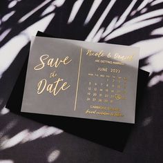 a silver and gold save the date card on top of a black tablecloth with palm leaves