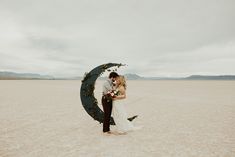 a bride and groom kissing in the middle of an empty field with a half moon