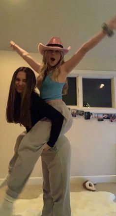 two young women are dancing in the living room with their arms spread out and one woman is wearing a cowboy hat