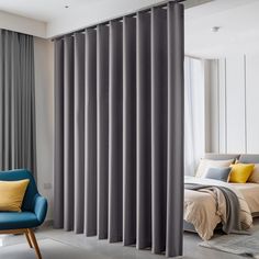 a blue chair sitting in front of a window next to a white bed and gray curtains