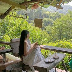 a woman sitting at a picnic table in front of a forest with trees and greenery