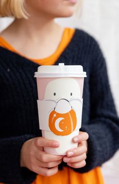 a woman holding a coffee cup with an orange and white design on the lid, in front of her