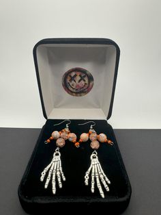 Embrace the spooky season with these Halloween dangling earrings that capture the essence of the holiday perfectly. Adorned with intricately designed skeleton hands and glass beads, each earring tells a whimsical tale of Halloween folklore. The lightweight yet eye-catching design ensures comfort while making a bold statement. Whether you're dressing up for a costume party or simply adding a touch of Halloween spirit to your ensemble, these earrings are the ideal accessory. Let these festive earrings be your playful companion throughout the Halloween season, adding a touch of macabre charm to any outfit. Celebrate the magic of Halloween with these unique, themed earrings that are sure to turn heads and spark conversations wherever you go. Drop Length: 3" Findings: Stainless Steel Handmade Metal Halloween Earrings, Handmade Metal Earrings For Halloween, Halloween Dangle Metal Earrings, Dangle Halloween Earrings, Spooky Pierced Earrings For Halloween, Metal Dangle Earrings For Halloween, Halloween Metal Drop Earrings, Adjustable Nickel-free Earrings For Halloween, Spooky Dangle Earrings