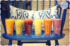 six glasses filled with different colored juices on a blue chair next to a pillow