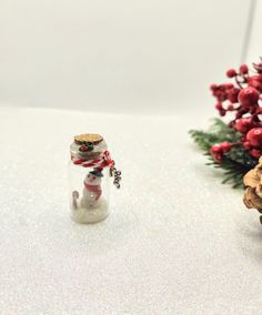 a small glass bottle with a snowman in it next to some pine cones and red berries