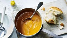 a bowl of soup with a spoon in it next to a piece of bread on a cutting board