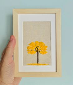 a hand is holding up a framed painting with a yellow tree on the front and bottom