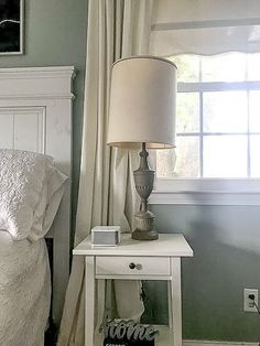 a bedroom with a bed, nightstand and lamp in front of a window that has white curtains