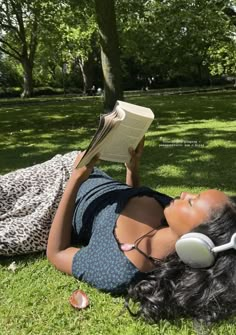 a woman laying in the grass reading a book and listening to headphones on her head