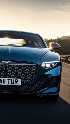 the front end of a blue car driving down a road at sunset with its headlights on