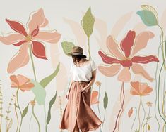 a woman standing in front of a wall with flowers and leaves on it, wearing a hat