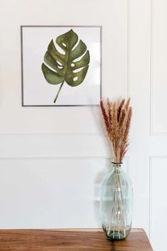 a glass vase with some plants in it on a wooden table next to a white wall