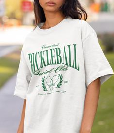 A fun graphic tee for the pickleballers! It's perfect for either the "social club" aesthetic or to wear when playing with your friends and teammates! For the "oversized" tee look, I recommend sizing up either 1 or 2 sizes from your typical day to day size! .: Made with 100% ring-spun cotton, a lightweight fabric (4.5 oz/yd² (153 g/m this unisex t-shirt feels like a bliss to wear all year round.  .: The classic fit with the crew neckline deliver a clean, versatile style that can match any occasion, whether it's formal or semi-formal.  .: All shirts feature a pearlized, tear-away label for total wearing comfort.  .: Made using ethically grown and harvested US cotton. Gildan is also a proud member of the US Cotton Trust Protocol ensuring ethical and sustainable means of production. This blank Pickleball Graphic Tee With Screen Print, Collegiate Crew Neck Tennis Tops, Casual Tennis T-shirt Relaxed Fit, Pickleball Sports Season Graphic Print T-shirt, Pickleball Sports Season Graphic T-shirt, Sporty T-shirt For Pickleball In Summer, Sporty T-shirt For Summer Pickleball, Sporty Short Sleeve T-shirt For Pickleball, Crew Neck Tennis T-shirt With Team Name