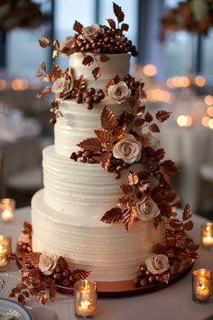 a white wedding cake with flowers and leaves on top is surrounded by candles in the background