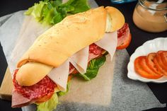 a sub sandwich with meat, cheese and tomatoes on a cutting board next to a bowl of dressing