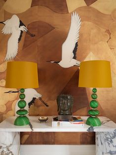 two lamps sitting on top of a white table next to a wall with birds painted on it