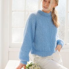 a woman wearing a blue sweater and white pants sitting on a table in front of a window