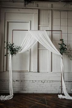 a white wedding arch decorated with flowers and greenery in front of a brick wall