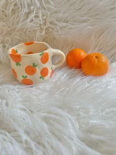 two oranges sitting next to a cup on a white fur covered surface, one is half peeled and the other half unpeeled