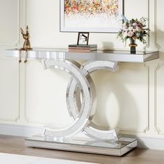 an elegant console table with a glass top and silver metal base, in front of a painting on the wall