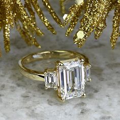 an emerald - cut diamond ring sits in front of a gold christmas tree ornament