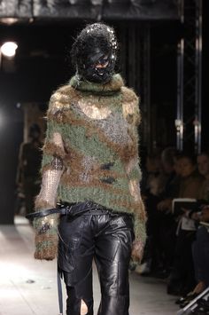 a model walks down the runway wearing black leather pants and a camouflage knitted sweater